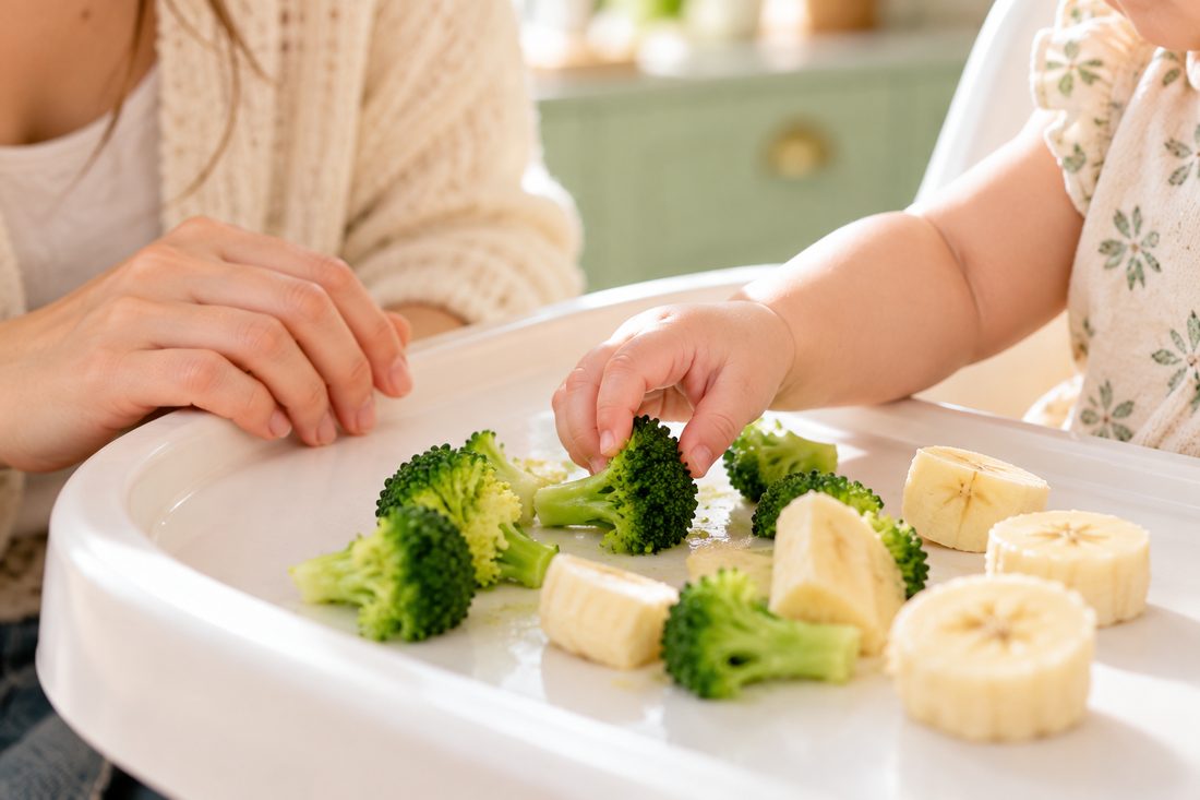 A baby picks up a soft broccoli floret with thumb and finger next to banana slices, while a parent's hand rests on the high chair tray